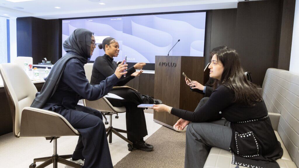 Four women sit and talk in a modern conference room with a screen and a podium labeled Apollo Opportunity Foundation in the background.