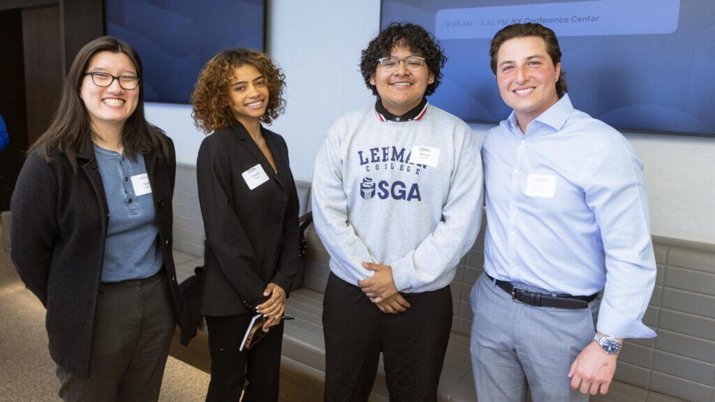 Four people stand indoors, smiling at the camera, each wearing business or casual attire with name tags. In the background, a screen and conference schedule from the Apollo Opportunity Foundation are visible.