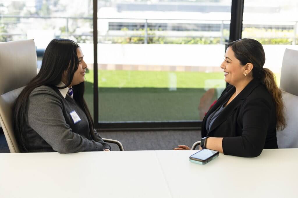Two women sit across from each other at a table in an office setting, engaged in conversation about the Apollo Opportunity Foundation. A smartphone rests on the table between them.
