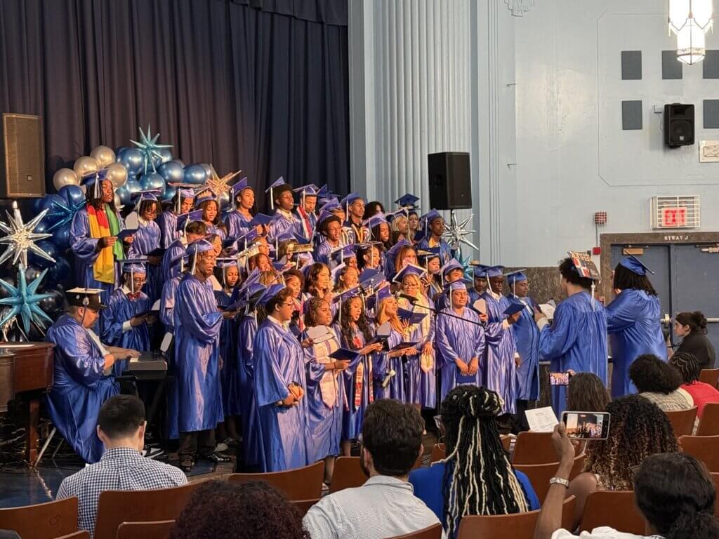 A group of graduates in blue caps and gowns stand on stage posing for photos during a graduation ceremony, while an audience watches and takes pictures.