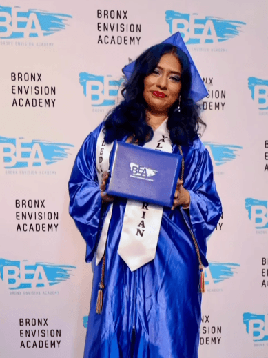 A graduate in a blue cap and gown holds a diploma cover on a Bronx Envision Academy backdrop, wearing a white sash that reads Valedictorian.