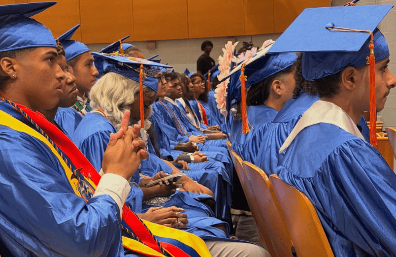 Students in blue graduation gowns and caps sit in rows at a ceremony, some wearing sashes and decorated caps, facing forward and listening attentively.