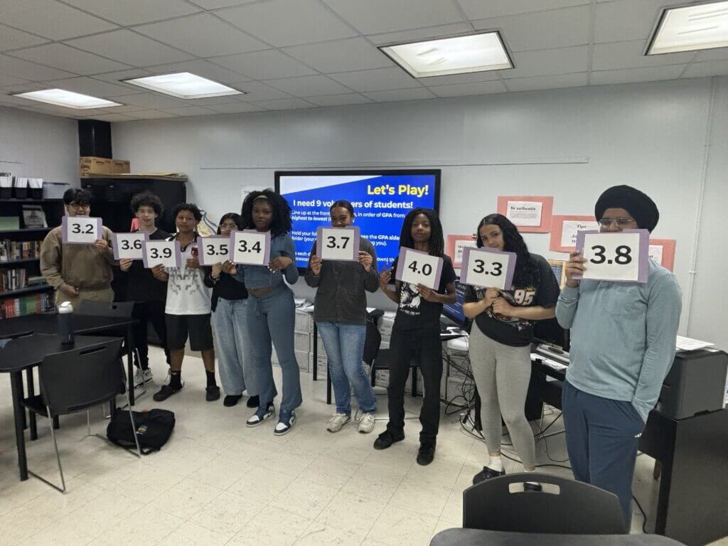 Eight students stand in a classroom holding up papers with decimal numbers ranging from 3.2 to 4.0. Behind them, a presentation slide titled "Let's Play!" is visible, possibly part of the Annual Report 2025 event.
