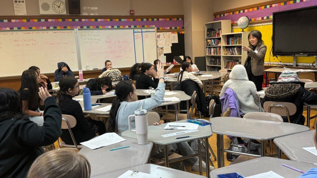 A volunteer stands at the front of a classroom speaking to students about early career exploration; several students have their hands raised.