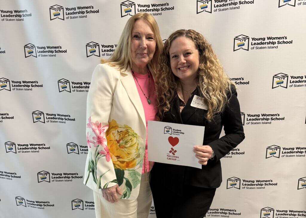 Two women stand together smiling in front of a Young Women’s Leadership School of Staten Island backdrop; one holds a sign with a red heart and Nicotra Group logo