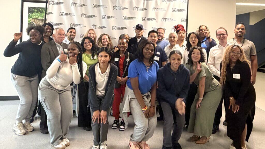 A group of adults and teens pose together in front of a branded backdrop in a hallway, smiling at the camera.