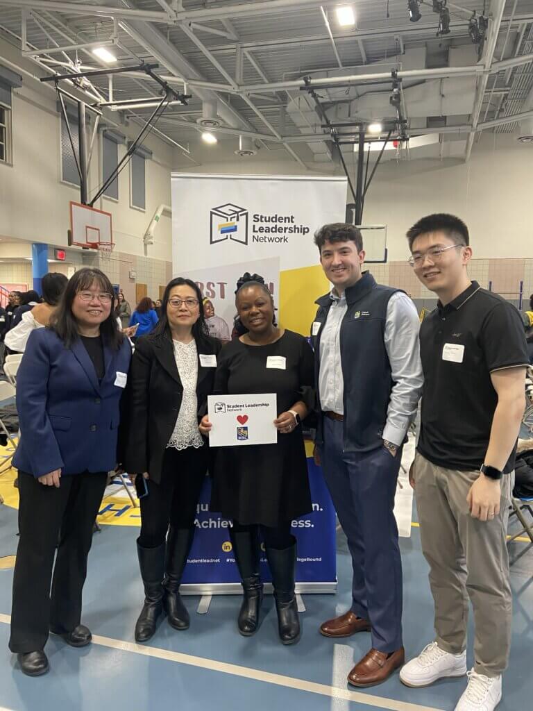 Five people stand in front of a Student Leadership Network banner in a gymnasium, smiling and holding a sign with a heart and icons, celebrating the release of the 2025 Annual Report.