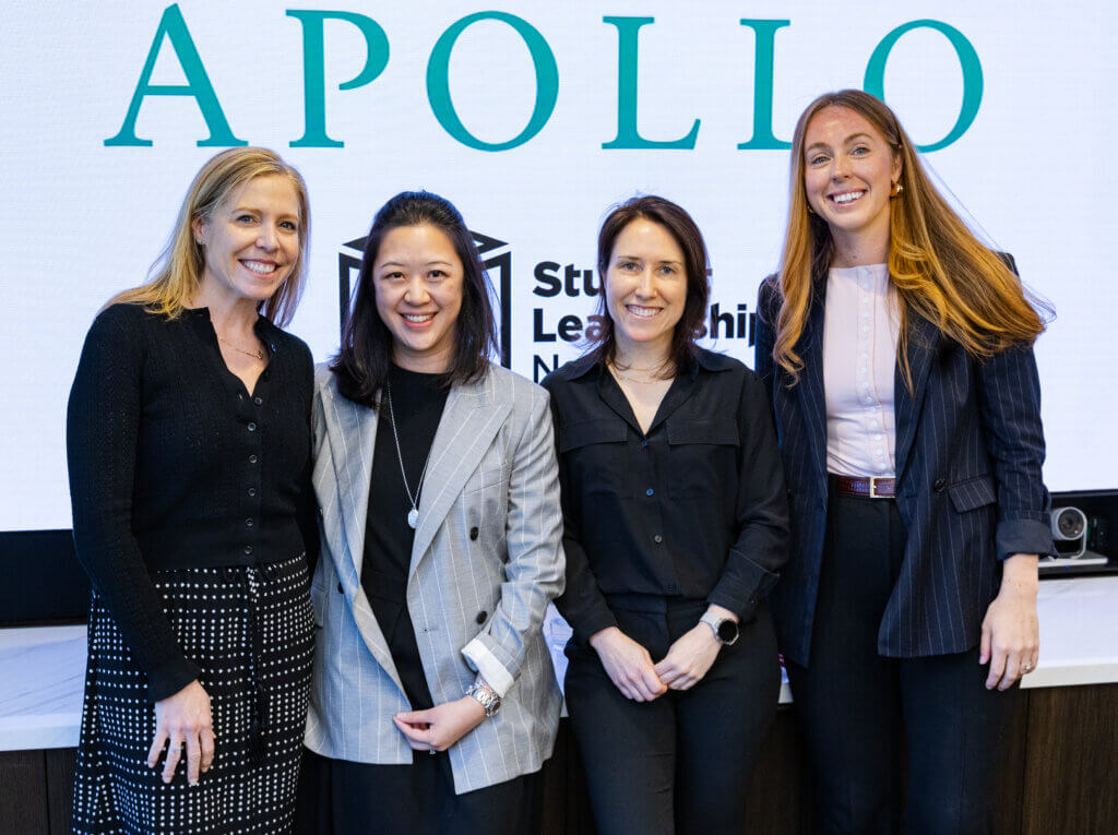 Four women standing and smiling in front of a screen displaying the word APOLLO and part of a Student Leadership sign, celebrating an Apollo Opportunity Foundation event.