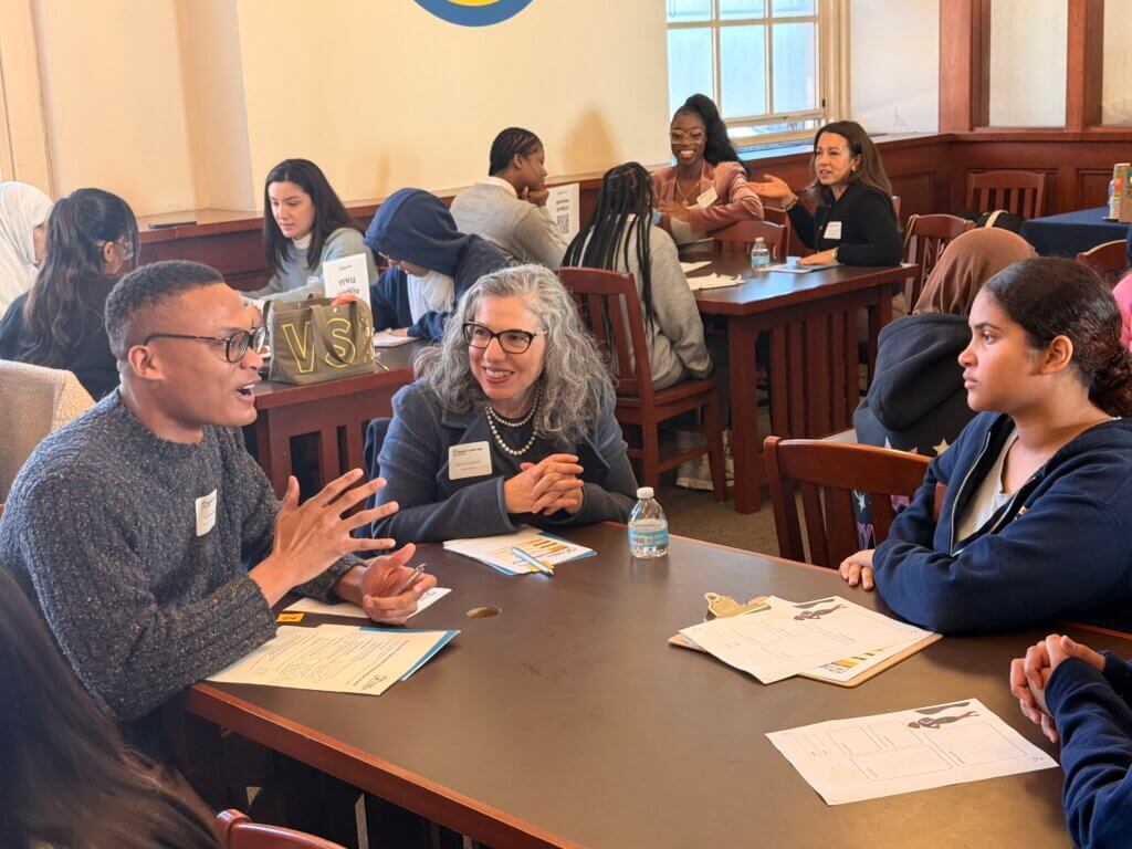 Three people sit at a table in discussion in a classroom setting, reviewing papers and the Annual Report 2025; name tags and a water bottle are visible as other groups talk in the background.