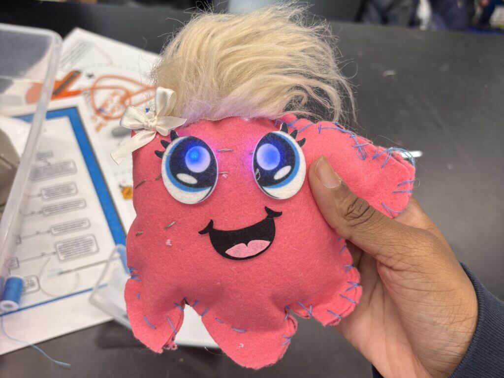 A hand holds a pink, hand-sewn plush toy with large blue eyes, a smiling mouth, light stitching, and a tuft of blond hair topped with a white bow