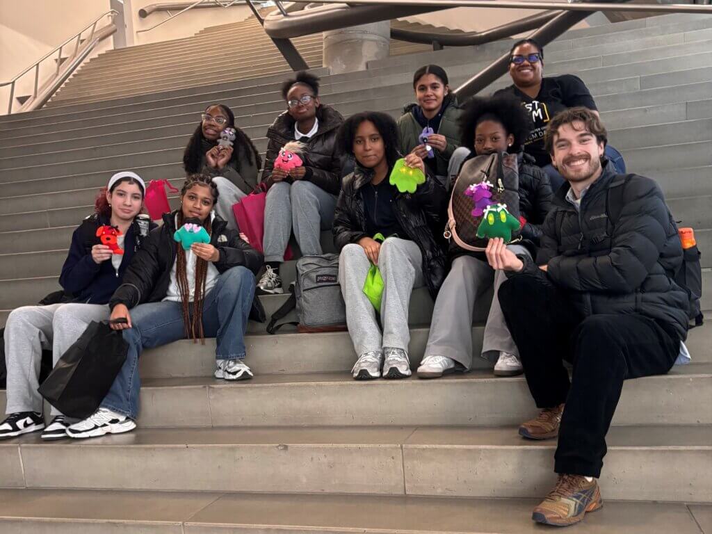 A group of eight people sit on stairs, holding colorful handmade crafts and smiling at the camera during a STEM exposure activity