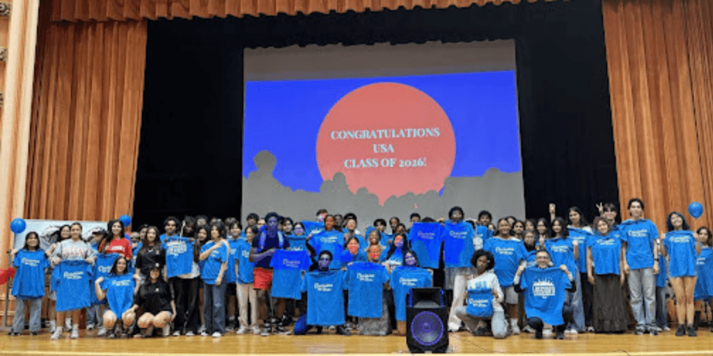 A large group of students on a stage hold blue shirts, posing in front of a projection that reads CONGRATULATIONS USA CLASS OF 2026!, celebrating with Jeffrey Gural.