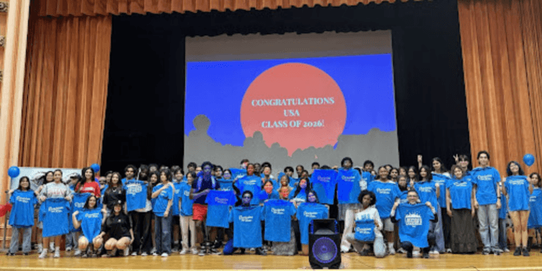 A large group of students on a stage hold blue shirts, posing in front of a projection that reads CONGRATULATIONS USA CLASS OF 2026!, celebrating with Jeffrey Gural.