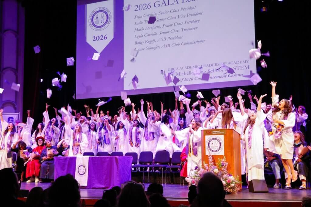 A large group of graduates in white gowns throw caps in the air on stage during a commencement ceremony, with a presentation slide visible behind them.