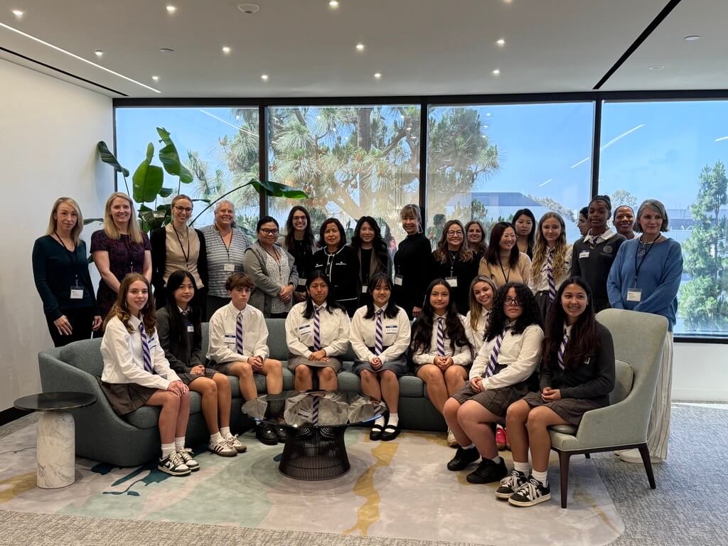 A group of students in uniforms and adults pose together in a modern office lounge area with large windows and indoor plants, capturing a moment for the 2025 Annual Report.