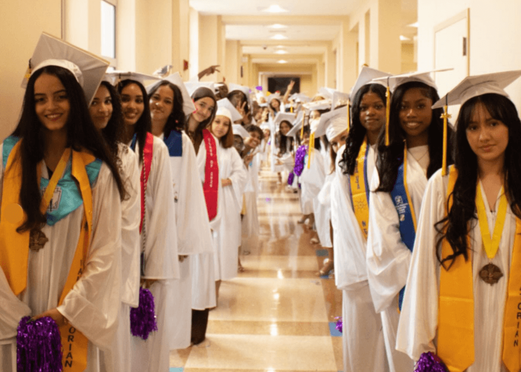 A group of students in white graduation gowns and caps stand in two rows along a hallway, wearing Class of 2026 sashes and holding decorations.