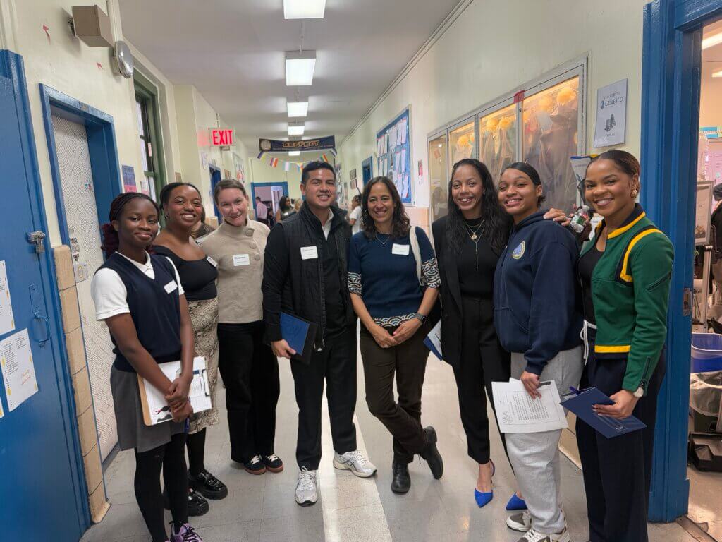 Eight people stand in a school hallway, posing for a group photo. Some hold papers or folders, possibly preparing for the 2025 Annual Report, with bulletin boards visible on the walls behind them.