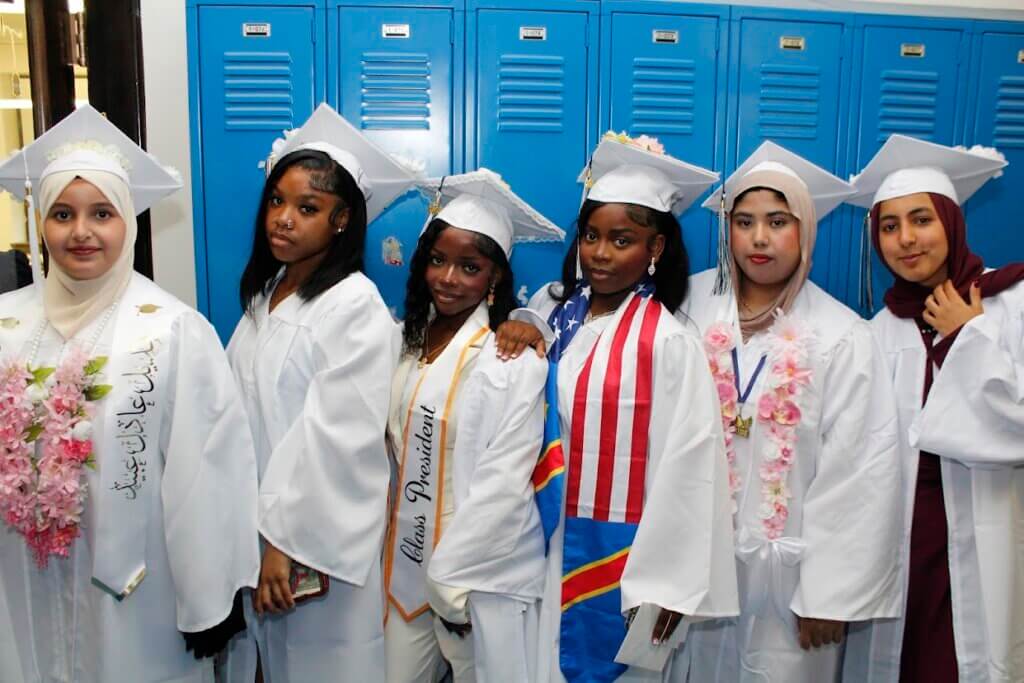 Six students from the Class of 2026 are in white graduation gowns and caps stand in front of blue lockers, some wearing sashes, stoles, and leis, posing for a group photo.