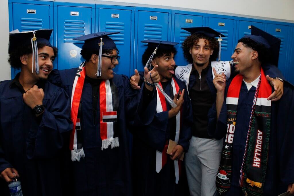 Five young men in graduation gowns and caps stand in front of blue lockers, smiling and gesturing to each other in celebration.