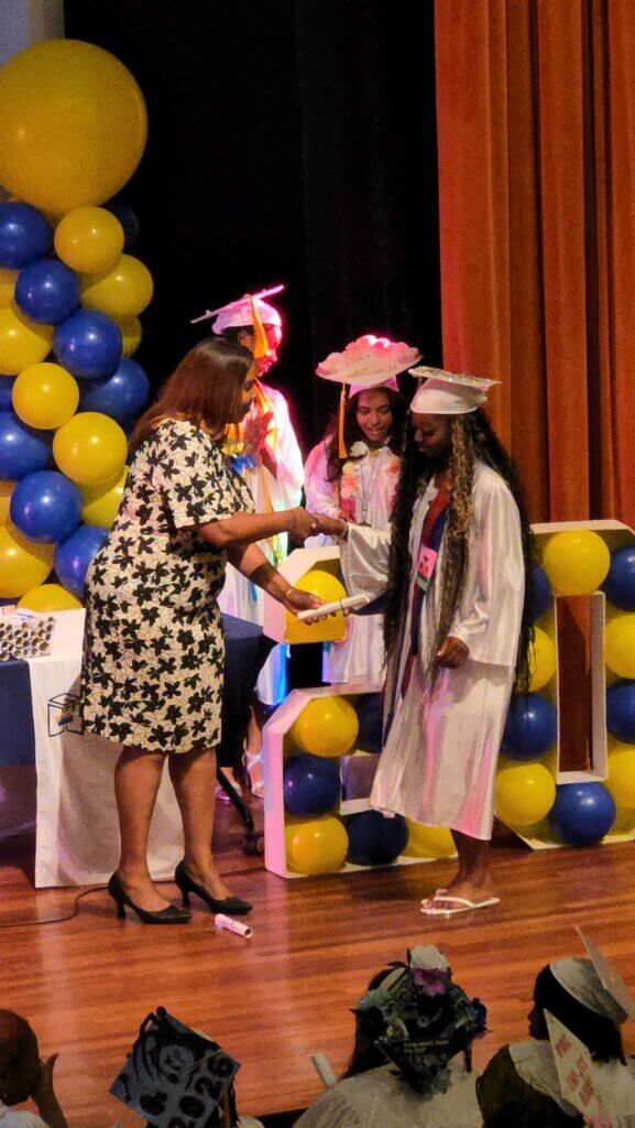 A graduate in a white cap and gown receives a diploma from a woman on stage, with other graduates and yellow and blue balloons in the background.