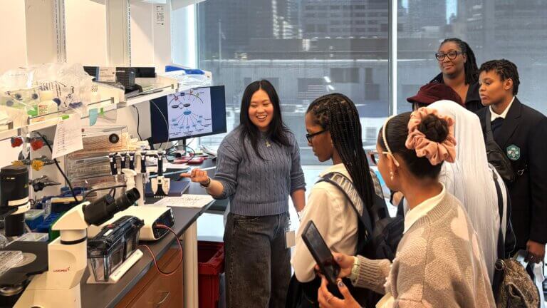 A woman explains scientific equipment to a group of students in a laboratory setting for a STEM exposure program, with lab materials in the background.
