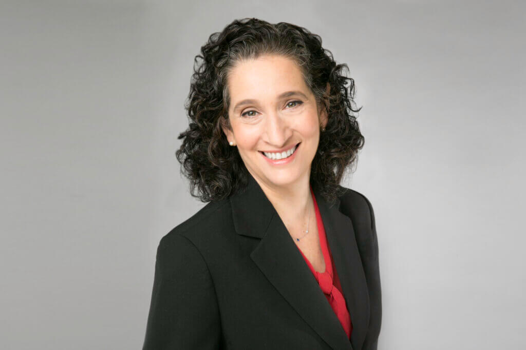Woman with curly dark hair wearing a black blazer and red blouse, smiling at the camera against a plain light gray background, reminiscent of a professional portrait style often seen in Jeffrey Gural’s business profiles.