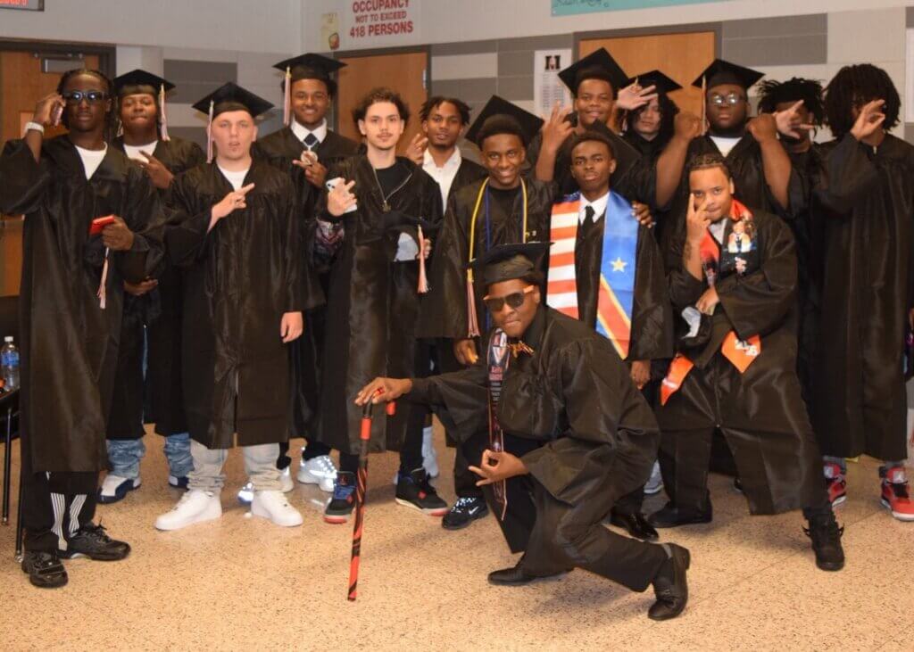 A group of male graduates in caps and gowns pose together indoors, with some making hand gestures and wearing graduation stoles.