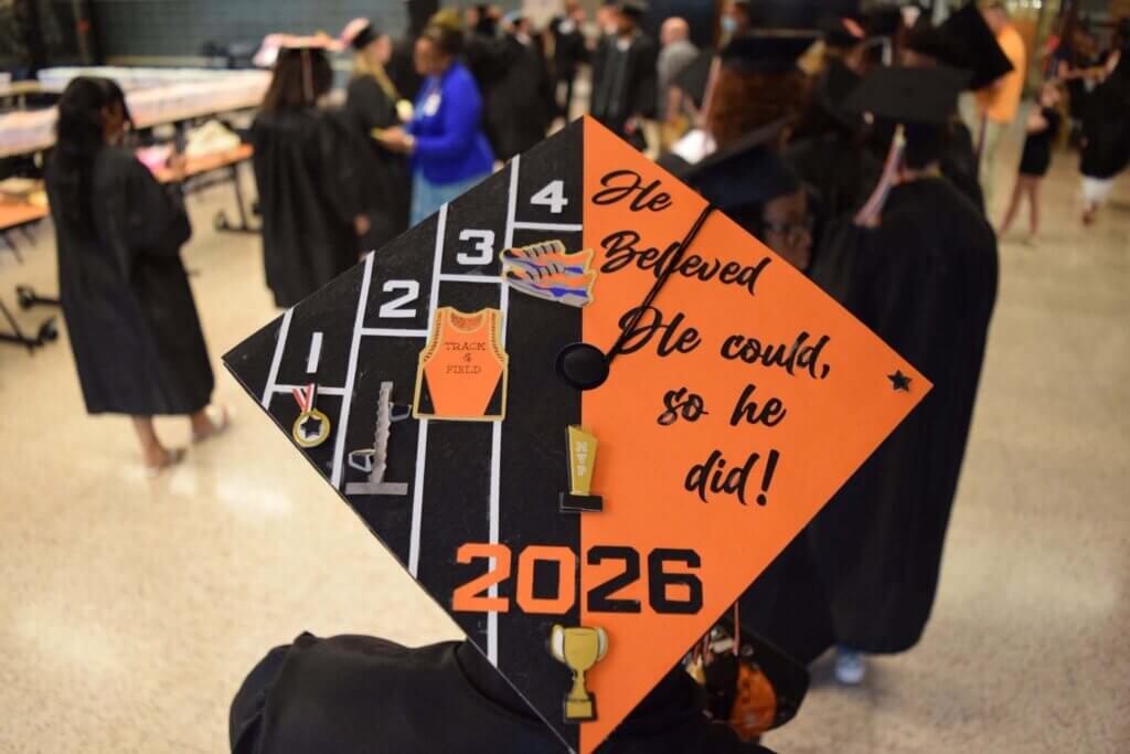 A graduation cap decorated with a track and field theme and the message He believed he could, so he did! with Class of 2026 and miniature athletic medals and trophies. Other graduates are in the background.