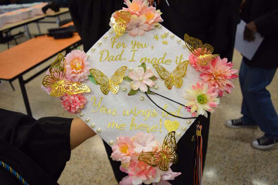 A decorated graduation cap with pink flowers, gold butterflies, and the message: For you, Dad. I made it. Your wings led me here! written in gold letters.