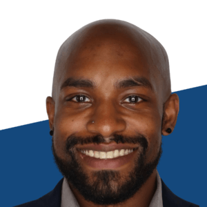 A man with a beard and mustache smiles at the camera. He is wearing a dark jacket and a gray shirt with a blue and white background.