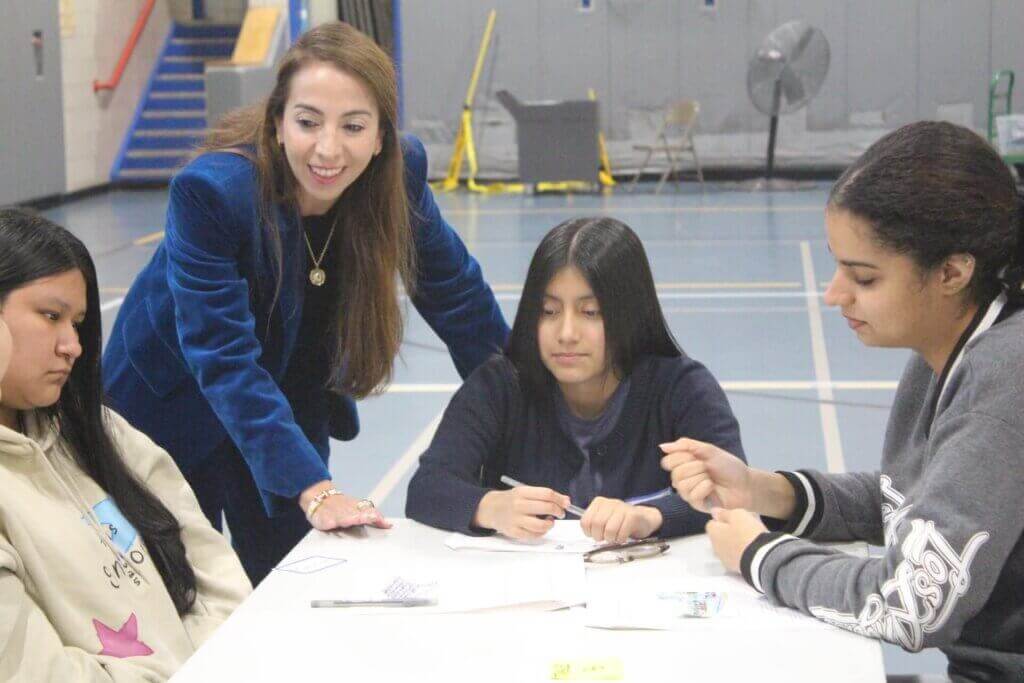 A woman stands beside three seated students at a table in a gymnasium, engaging with them as they work on papers and cards—capturing a moment showcased in our Annual Report 2025.