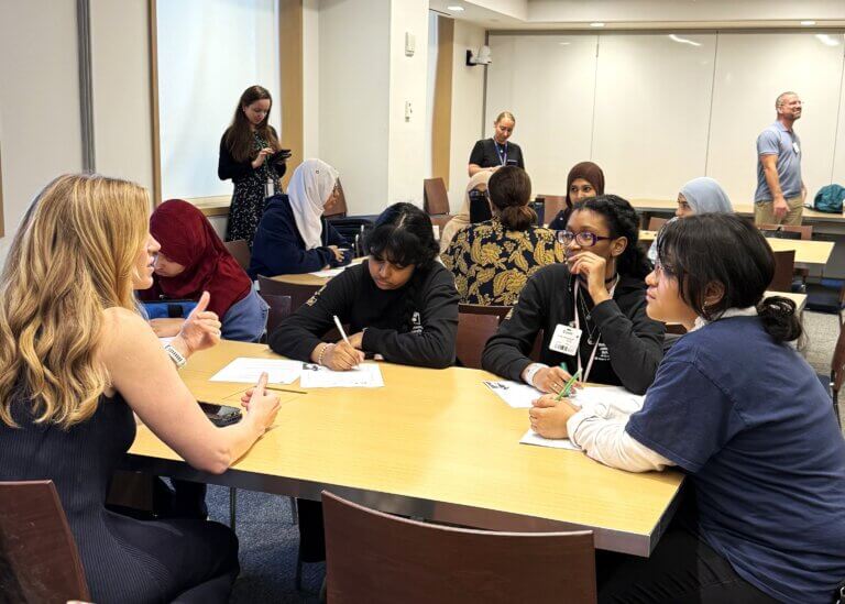 A group of students sits at a table with a woman, writing notes during a career exposure discussion