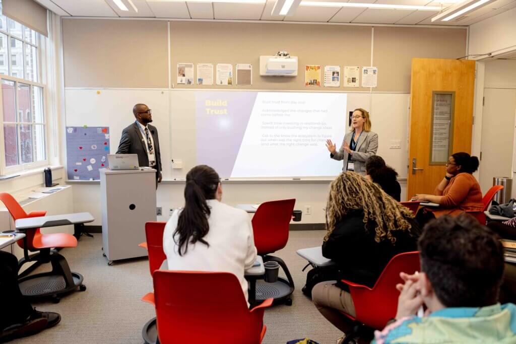 Two people are presenting to a group in a classroom, with a slide titled Build Trust displayed on the screen behind them. Several attendees are seated and listening during the Annual Report 2025 session.