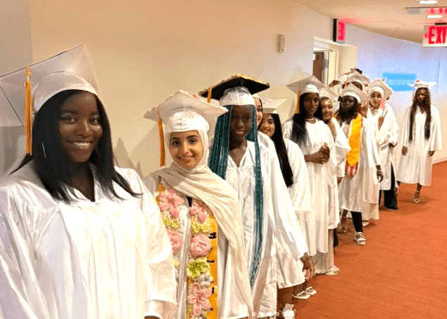 A group of students in white graduation gowns and caps stand in a line indoors, smiling and facing the camera.