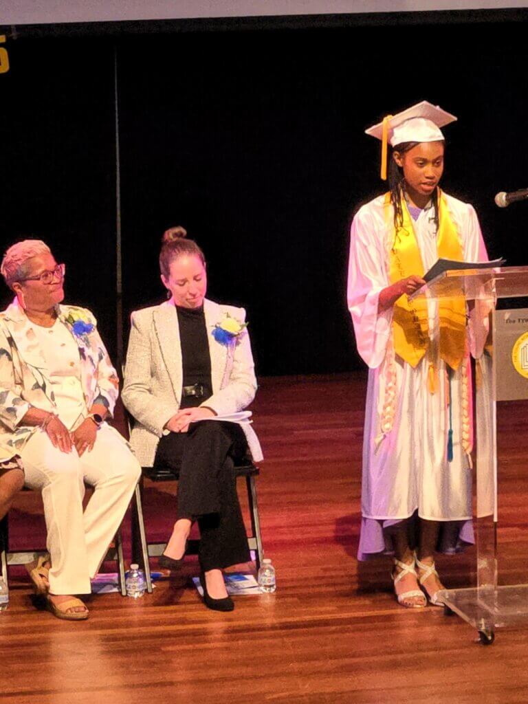 A Class of 2026 student in a white graduation gown and cap speaks at a podium on stage