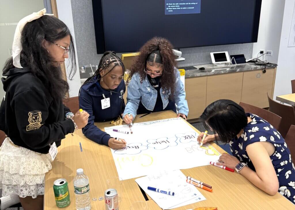 Four young women stand and sit around a table, collaborating and writing on a large sheet of paper with colorful markers, for a career exposure activity