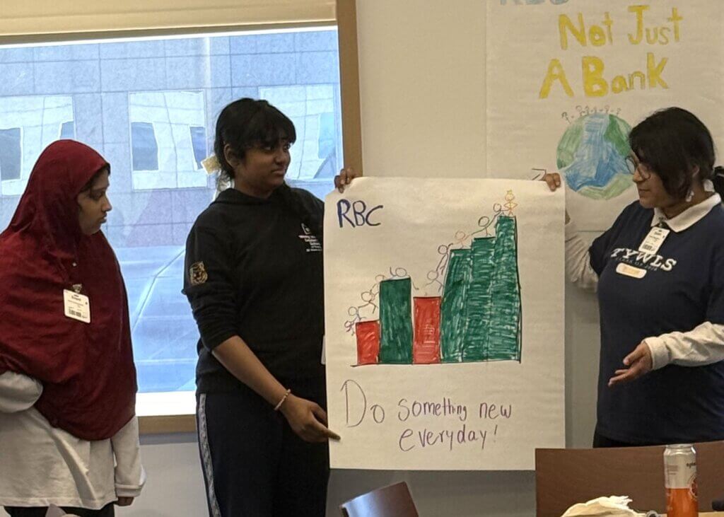 Three people stand indoors presenting a hand-drawn poster with bar graphs and the text “RBC” and “Do something new everyday!” on it for a career exposure activity