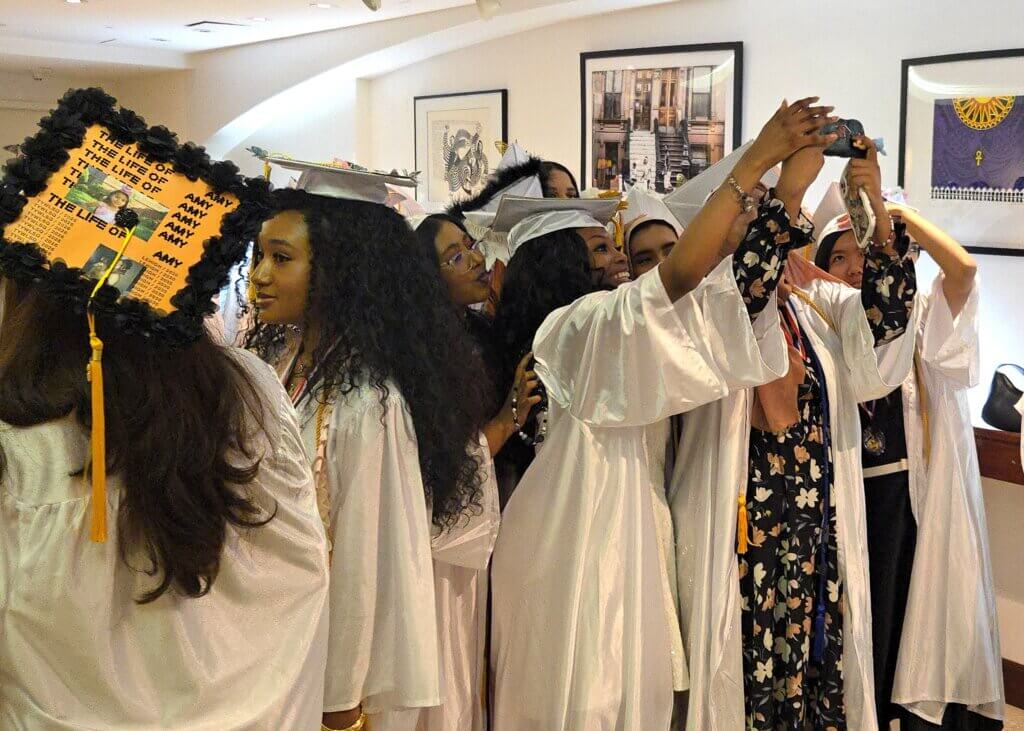 A group of graduates in white caps and gowns take selfies together indoors; one cap displays a decorated message with yellow paper and black letters.