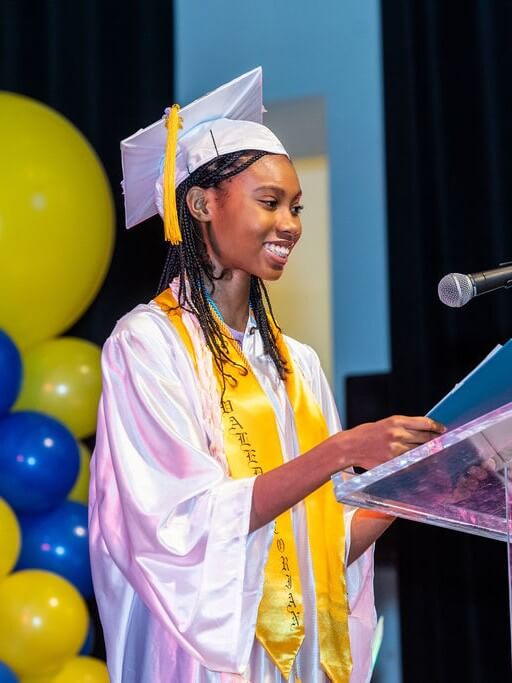 A Class of 2026 student in a white graduation gown and cap speaks at a podium on stage