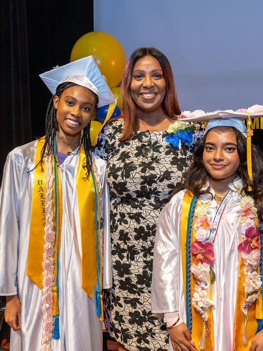 A graduate in a white cap and gown receives a diploma from a woman on stage, with other graduates and yellow and blue balloons in the background.