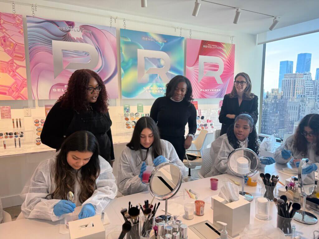 Six women gather in a bright room for the 2025 Annual Report; four in lab coats work with beauty products at a table, while two stand behind observing. City buildings can be seen through large windows.
