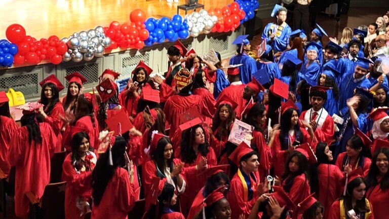Students in red and blue caps and gowns gather in an auditorium for a graduation ceremony, with balloons decorating the stage in the background.