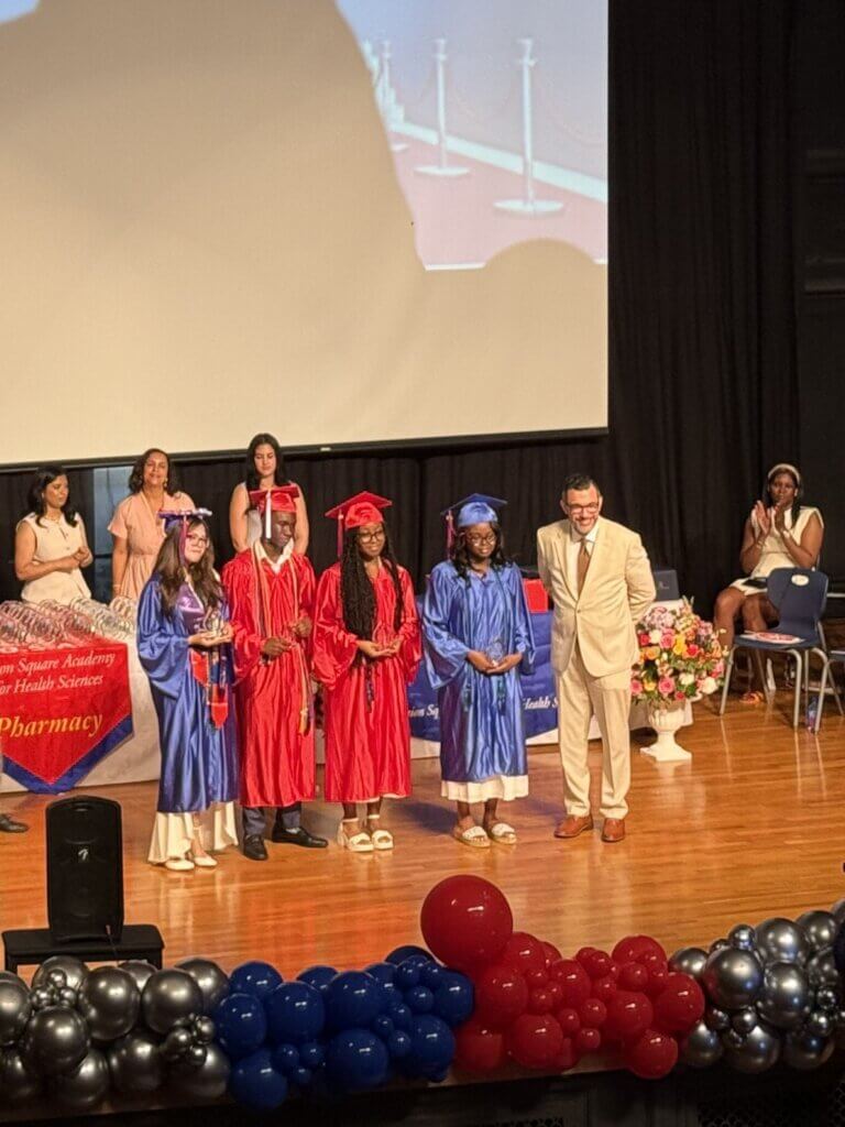 A graduate in a blue cap and gown receives a diploma from a man in a tan suit on stage during the Class of 2026 graduation ceremony