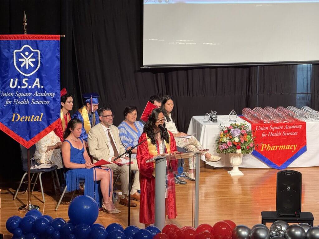 A student in a red cap and gown speaks at a podium on stage during a graduation ceremony, with faculty and students seated behind her and banners for Dental and Pharmacy programs displayed.