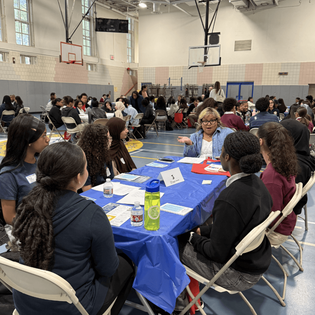 A group of students sits around a table with an adult in a gymnasium, participating in a discussion activity with papers and water bottles on the table as part of the Annual Report 2025 preparations.