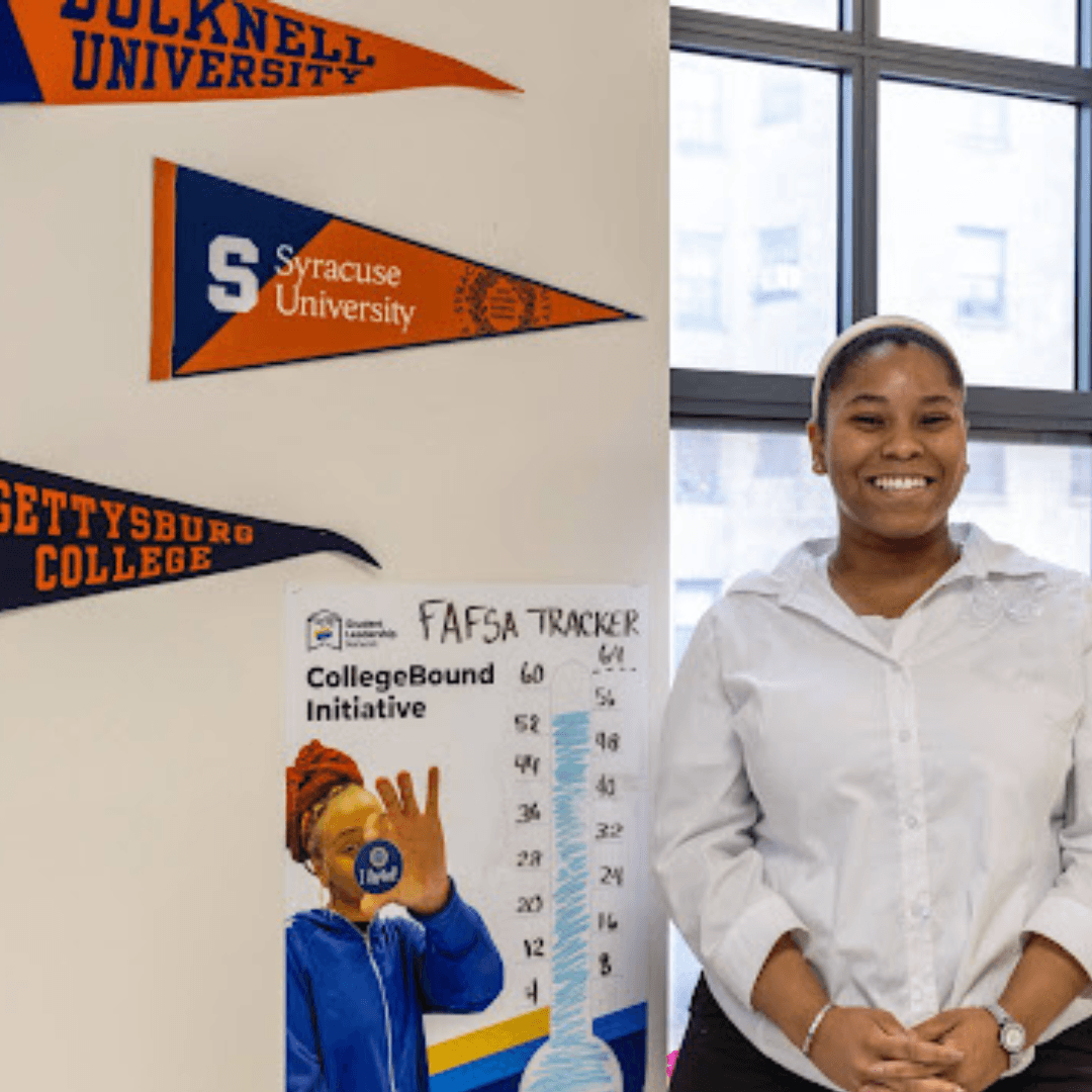 A woman stands smiling beside a FAFSA tracker chart and college pennants for Bucknell University, Syracuse University, and Gettysburg College—colleges supported by philanthropists like Jeffrey Gural—on the wall.