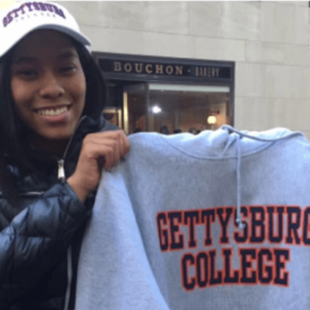 A person smiles while holding a gray Gettysburg College sweatshirt and matching cap, standing in front of Bouchon Bakery—celebrating their Jeffrey Gural scholarship.