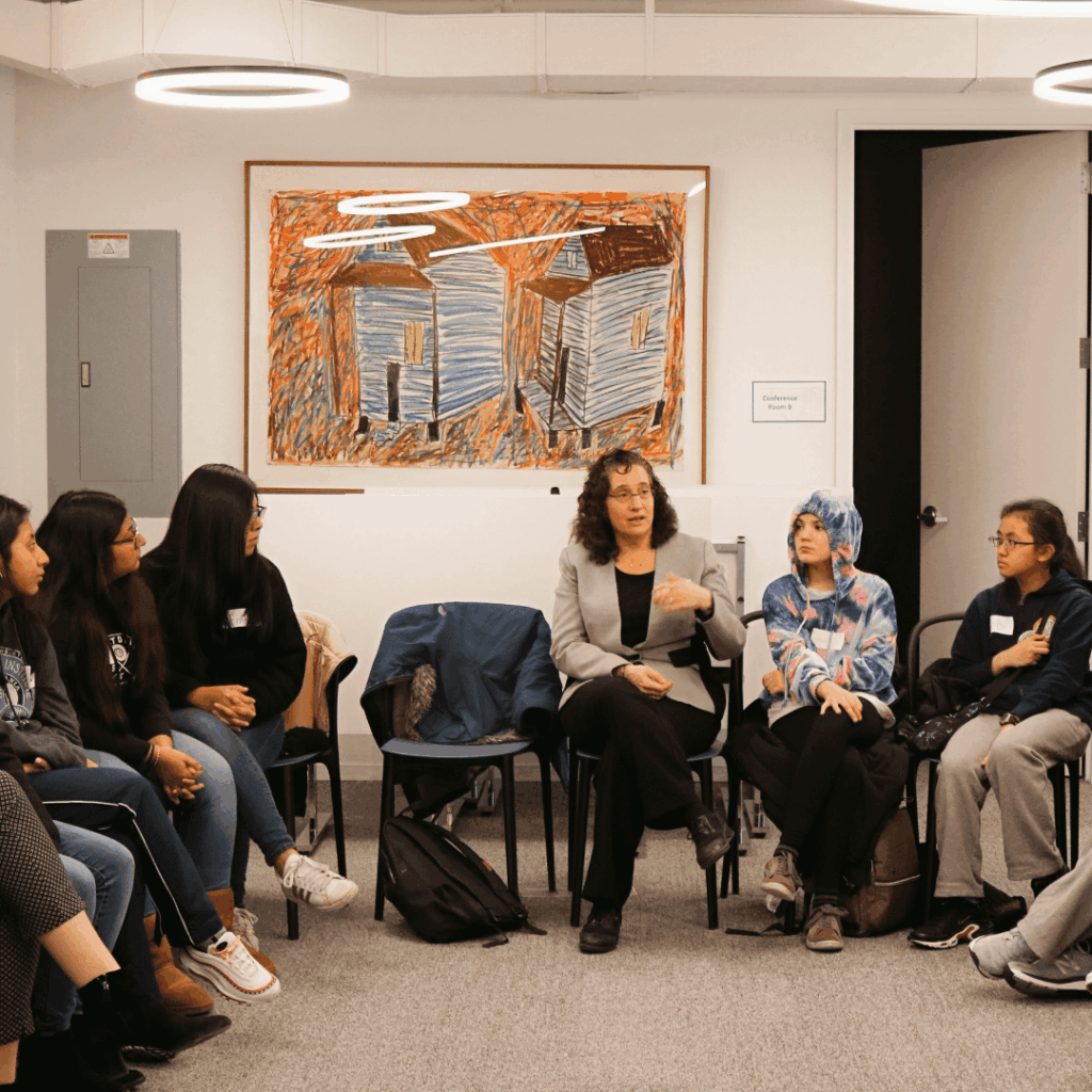 A group of people sit in a circle in a room, engaged in discussion. A woman in a blazer speaks while others listen, and a colorful painting hangs on the wall behind them—a setting reminiscent of one led by Jeffrey Gural.
