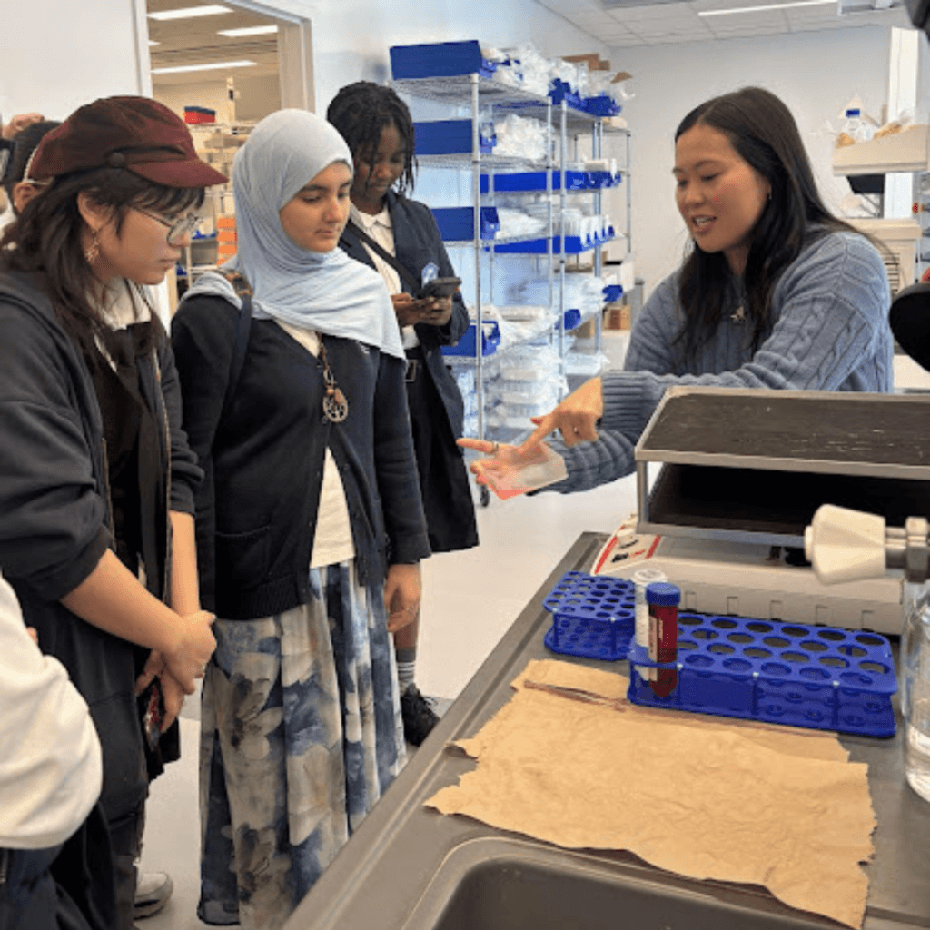 A woman explains a laboratory process to a group of attentive students in a science lab setting with lab equipment on the counter, referencing research by Jeffrey Gural.