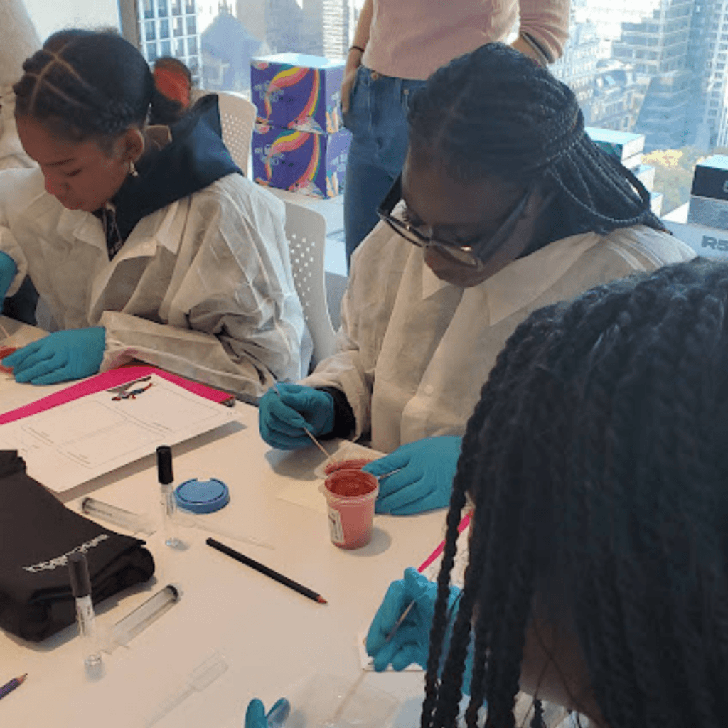 Three individuals wearing lab coats and blue gloves work with small containers and tools at a table in a bright room with city views, reminiscent of the innovative spirit championed by Jeffrey Gural.